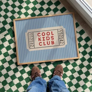 Puede incluir: Un estampado enmarcado con un fondo azul claro y rayas blancas verticales. El estampado presenta un diseño de boleto con las palabras "COOL KIDS CLUB" en rojo. El marco es de madera clara. La imagen está sobre una alfombra a cuadros verdes y blancos.
