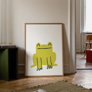 May include: A framed art print featuring a cheerful, cartoon-style frog in bright yellow. The frog has a simple design with black outlines and a happy expression. The artwork is set against a white background within a wooden frame.