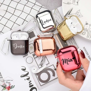 May include: Five personalised Airpod cases in different colours. The cases are silver, gold, black, rose gold, and red. Each case has a different name written on it: Lauren, Jackie, Mia.