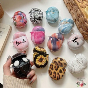 May include: Assortment of colorful Airpod cases with various patterns, including marble, zebra, leopard, sunflower, and floral designs. Some cases have names written on them, such as "Norah" and "Lea".