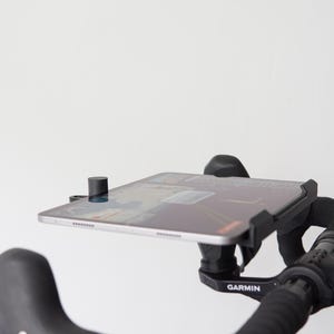 Op de afbeelding: Een zwarte tablethouder bevestigd aan een fietsstuur met een Garmin stuurpenhouder. Het apparaat houdt een dunne tablet horizontaal vast, voorzien van een zwart frame en een kleine cilindrische knop linksboven op het scherm.