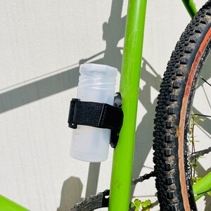Könnte beinhalten: Eine durchsichtige Plastikwasserflasche ist mit einem schwarzen Band an einem grünen Fahrradrahmen befestigt. Das Fahrrad hat einen braunen Reifen mit einem Profil.