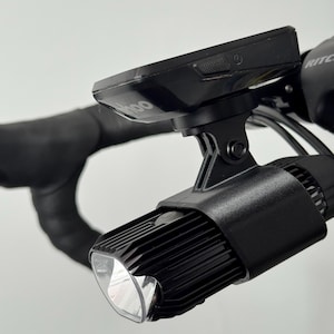 Peut inclure: Feu avant de vélo noir et ordinateur de cyclisme montés sur le guidon. La lumière a une forme rectangulaire avec une lentille claire. L'ordinateur de cyclisme est noir avec la marque "Wahoo" visible. Le guidon est recouvert de ruban adhésif noir.