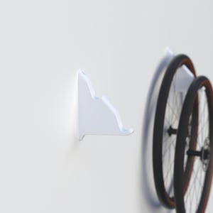 May include: White plastic bike wheel hook mounted on a white wall. Two black bike wheels are leaning against the hook.