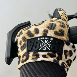 Peut inclure: Un gant de sport à motif léopard avec un écusson en caoutchouc noir portant l'inscription "LRGA" et "CULT OF SHRED" en blanc. Le gant possède un poignet côtelé noir et est placé sur une poignée de guidon noire avec un levier de frein noir.