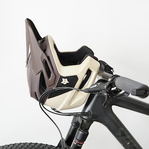 Könnte beinhalten: Ein zweifarbiger Mountainbike-Helm in Braun und Creme mit großem Visier und Fox-Logo an der Seite, der auf dem Lenker eines schwarzen Mountainbikes liegt. Der Helm verfügt über mehrere Belüftungsöffnungen und schwarze Innenpolster.