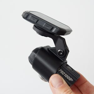 May include: A black electronic device with a screen mounted on an adjustable arm, featuring the text PRO1000+ printed on the side. The unit has a rugged, textured casing and a hinge mechanism that allows for positioning the display screen.