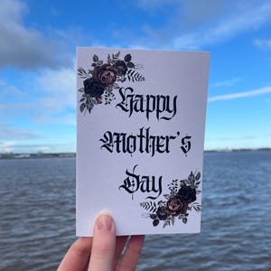 May include: A white card with black gothic lettering that reads "Happy Mother's Day". The card is decorated with black and pink floral illustrations.