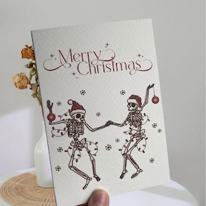 May include: A Christmas card featuring two dancing skeletons wearing Santa hats and wrapped in Christmas lights. The card has the words "Merry Christmas" in a decorative font, with snowflakes scattered around the skeletons.