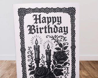May include: A black and white birthday card with the words "Happy Birthday" in a gothic font. The card features illustrations of lit candles, roses, and decorative flourishes within a detailed border.