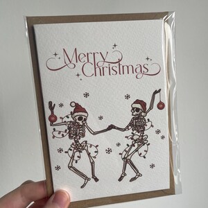 May include: A Christmas card featuring two dancing skeletons wearing Santa hats and string lights. The words "Merry Christmas" are written in a decorative font above the skeletons. The card is in a clear plastic sleeve.