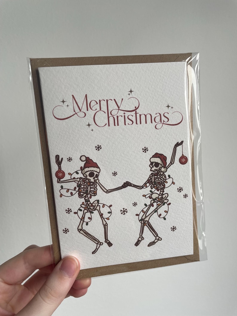 May include: A Christmas card featuring two dancing skeletons wearing Santa hats and string lights. The words "Merry Christmas" are written in a decorative font above the skeletons. The card is in a clear plastic sleeve.
