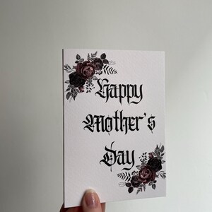 May include: A white card with black gothic lettering that reads "Happy Mother's Day". The card is decorated with black and pink floral illustrations.