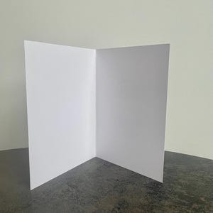 May include: An open, blank white greeting card, standing upright on a dark gray surface. The card is folded in half, revealing a clean, empty interior. The background is a plain, light-colored wall. The card is ready for a personalized message.