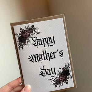 May include: A white card with a black and white gothic font that says "Happy Mother's Day". The card is decorated with black and burgundy watercolor roses and leaves.