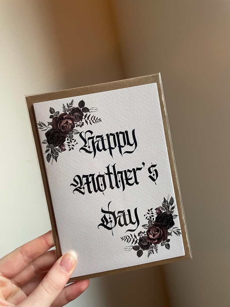 May include: A white card with a black and white gothic font that says "Happy Mother's Day". The card is decorated with black and burgundy watercolor roses and leaves.