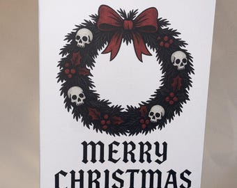 May include: A white greeting card with a black wreath design featuring skulls and a red bow. The words "MERRY CHRISTMAS" are printed in a gothic font below the wreath. The card is held by a person.