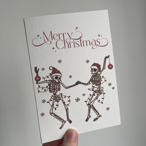 May include: A white greeting card with the words "Merry Christmas" in burgundy script. The card features illustrations of two skeletons dancing, adorned with Santa hats, Christmas lights, and ornaments. Snowflakes are scattered around the skeletons.