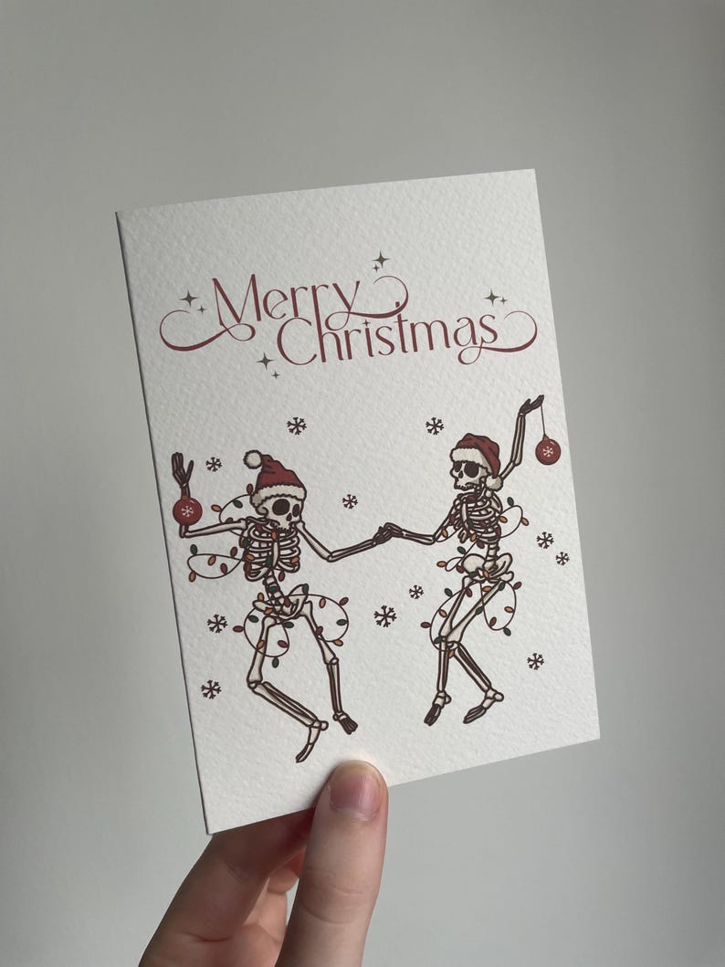 May include: A white greeting card with the words "Merry Christmas" in burgundy script. The card features illustrations of two skeletons dancing, adorned with Santa hats, Christmas lights, and ornaments. Snowflakes are scattered around the skeletons.