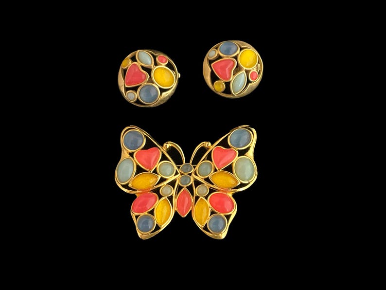 May include: A gold butterfly brooch with red, yellow, and blue enamel accents. The brooch is made of metal and has a detailed design. The butterfly has a pair of matching earrings with the same design.