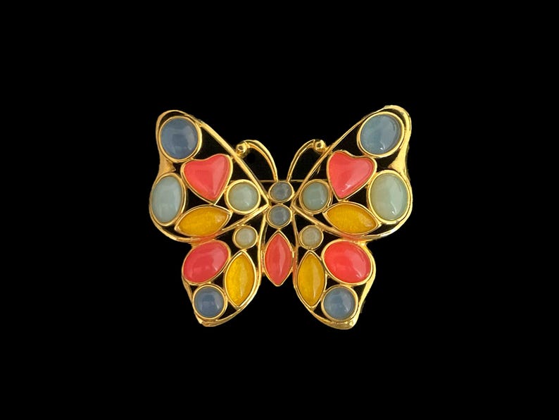 May include: A gold-tone butterfly brooch with colorful enamel accents. The butterfly has blue, yellow, pink, and red enamel details.