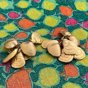 May include: Gold-toned seashell earrings. Each earring features a cluster of detailed clam shell charms. The earrings are displayed on a colorful fabric with a fish pattern in shades of green, orange, yellow, and pink.