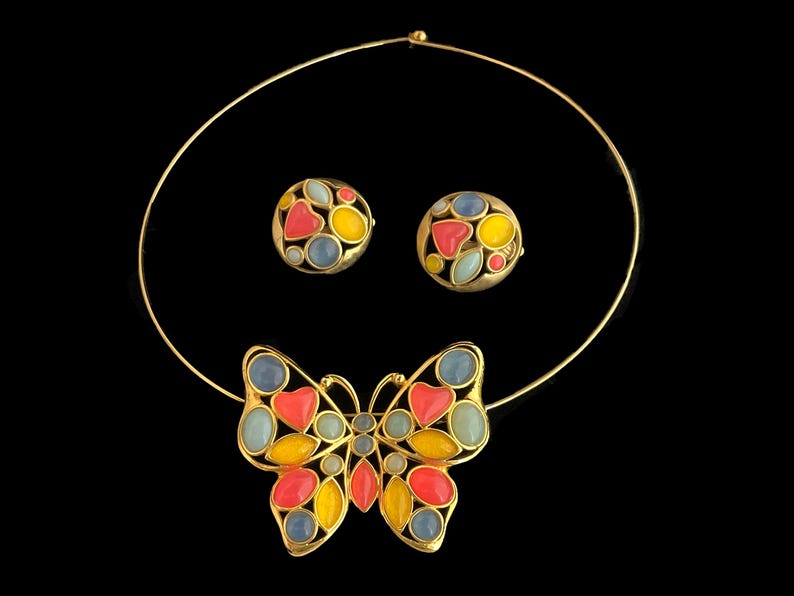 May include: A gold necklace with a butterfly pendant. The butterfly is made of gold and has red, yellow, blue, and green enamel. The necklace also comes with a pair of matching earrings.