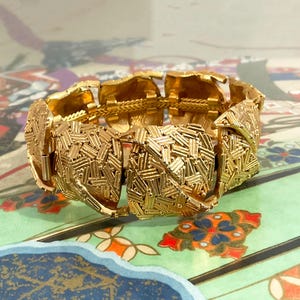 Può includere: Un braccialetto dorato con un design strutturato e intrecciato. Il braccialetto è composto da maglie rettangolari e presenta una chiusura a fermaglio. Il motivo intricato conferisce al braccialetto un aspetto vintage o antico. Il braccialetto è esposto su una superficie a motivi.