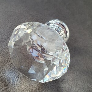 May include: A clear, faceted glass cabinet knob with a multi-sided geometric shape. The knob features a polished silver-tone metal base that connects to the back, providing a reflective surface that catches light through the transparent glass structure.