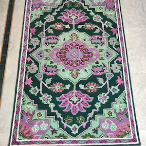 Puede incluir: Una alfombra de color verde, rosa y blanco con un diseño floral. La alfombra tiene un borde blanco y un borde interior verde y rosa.