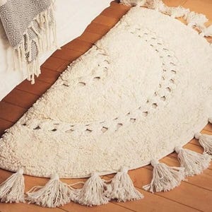 Può includere: Un tappeto a forma di mezza luna di colore crema con un design a uncinetto e bordi a frangia. Il tappeto presenta un motivo delicato e intricato ed è realizzato in un materiale morbido e soffice.