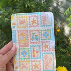 May include: A rectangular sticker sheet with a repeating pattern of postage stamp-style illustrations. The stamps feature images of a beach scene, scooter, flowers, wine, and sea creatures in pastel colors. The word "AMALFI" is printed on one of the stamps.