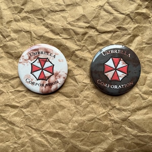 Op de afbeelding: Twee ronde buttons met de tekst "Umbrella Corporation" rond een rood-wit embleem. Eén button is wit met een roodbruine achtergrond, de andere is donkerbruin.