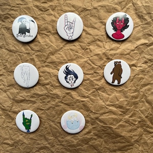 May include: A collection of nine white round buttons with various illustrations. Designs include a grim reaper, a hand making a rock and roll gesture, a devil, a cat-like creature, a woman with multiple eyes, a bear, and a baby in a bowl.