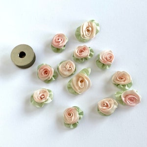 Op de afbeelding: Een verzameling van 12 kleine, roze en groene stoffen rozenversieringen. De rozen zijn gemaakt van satijnen lint en hebben een gelaagde, gerimpelde uitstraling.