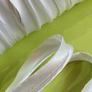 May include: Close-up of white satin ribbon with a stitched edge, arranged on a bright green background. The ribbon is folded and curved, creating a textured appearance. Ideal for crafting and sewing projects.