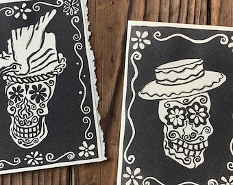 May include: Two black and white Day of the Dead art prints. Each features a sugar skull design with a hat, framed by a white floral border. The prints are on a textured surface.