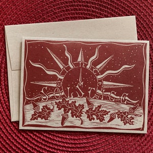 Pode incluir: Um cartão vermelho com um desenho de sol branco e as palavras "Solstice" impressas nele. O sol tem um mostrador de relógio com algarismos romanos. Há cinco bolotas brancas na parte inferior do cartão.