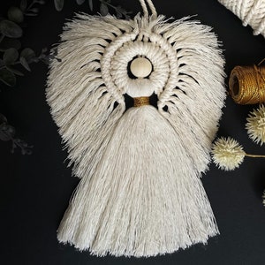 May include: White macrame angel ornament with a wooden bead and gold accents. The angel has a long tassel and is hanging from a white cord.