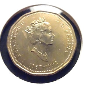 May include: A gold-coloured Canadian one-dollar coin featuring a profile portrait of Queen Elizabeth II. The text ELIZABETH II and D.G. REGINA is inscribed along the edge, with the commemorative dates 1867-1992 printed at the bottom of the coin.