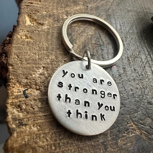 May include: A silver keyring with a circular pendant. The pendant is stamped with the words "You are stronger than you think" in a black, sans-serif font. The keyring is attached to a silver key ring.