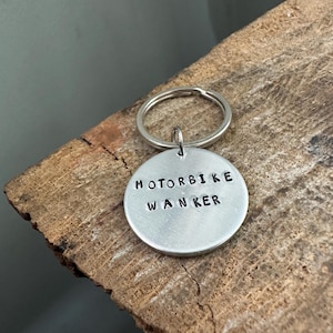 May include: A circular silver-coloured metal keyring featuring the stamped black text MOTORBIKE WANKER. The round metal tag is attached to a standard silver-coloured split ring, providing a simple and functional design for holding keys securely.