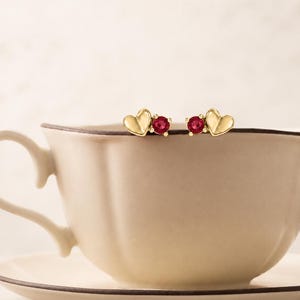 May include: A pair of gold stud earrings, each featuring a small heart-shaped element paired with a round, faceted red gemstone set in a four-prong gold setting. The earrings are positioned on the rim of a light-colored ceramic teacup.