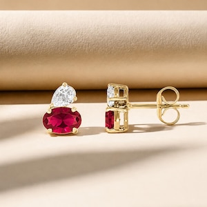 May include: A pair of gold stud earrings featuring a vibrant oval-cut red gemstone paired with a smaller pear-cut clear white gemstone. The earrings are secured with gold posts and butterfly backs, showcasing a polished metal setting.