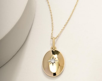 May include: A gold-colored pendant necklace featuring an oval pendant with a starburst design and a small diamond at the center. The necklace has a delicate chain and a simple clasp. The background is a soft, neutral color.