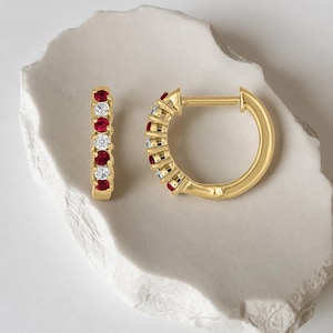 May include: A pair of gold hoop earrings featuring an alternating pattern of round-cut red rubies and clear white diamonds. The earrings have a hinged post closure mechanism and are designed with a polished gold finish along the circular band.