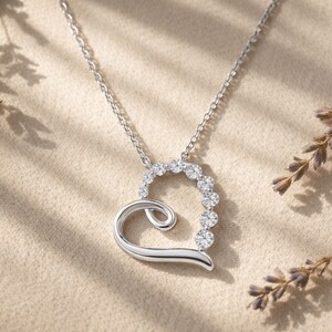 May include: A silver heart-shaped pendant necklace with a chain. The pendant features a curved design with a row of round, sparkling gemstones along the top curve. The necklace is displayed on a neutral-toned surface.