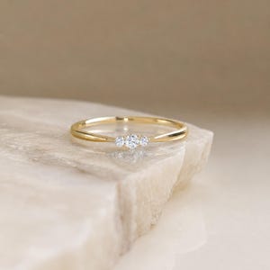 May include: A thin gold band ring featuring three small, round, clear, faceted stones set horizontally in the center. The metal has a polished, reflective finish, and the stones are arranged in a row, creating a delicate and simple design.
