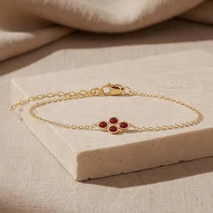 May include: A delicate gold chain bracelet featuring a central clover-shaped charm composed of four small, round, deep red gemstones. The bracelet includes a thin link chain and a secure lobster claw clasp with an adjustable extender chain.
