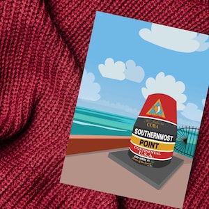 Op de afbeelding: Een ansichtkaart met het zuidelijkste punt van het continentale vasteland van de VS in Key West, Florida. De illustratie toont de iconische boei tegen een achtergrond van de oceaan, de lucht en wolken. De ansichtkaart ligt op een rode gebreide achtergrond.